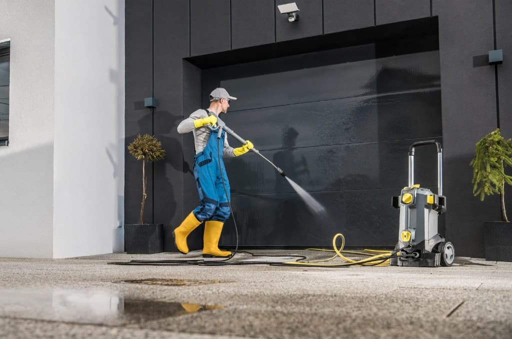 Worker in blue overalls pressure washing a large dark garage door and wet driveway
