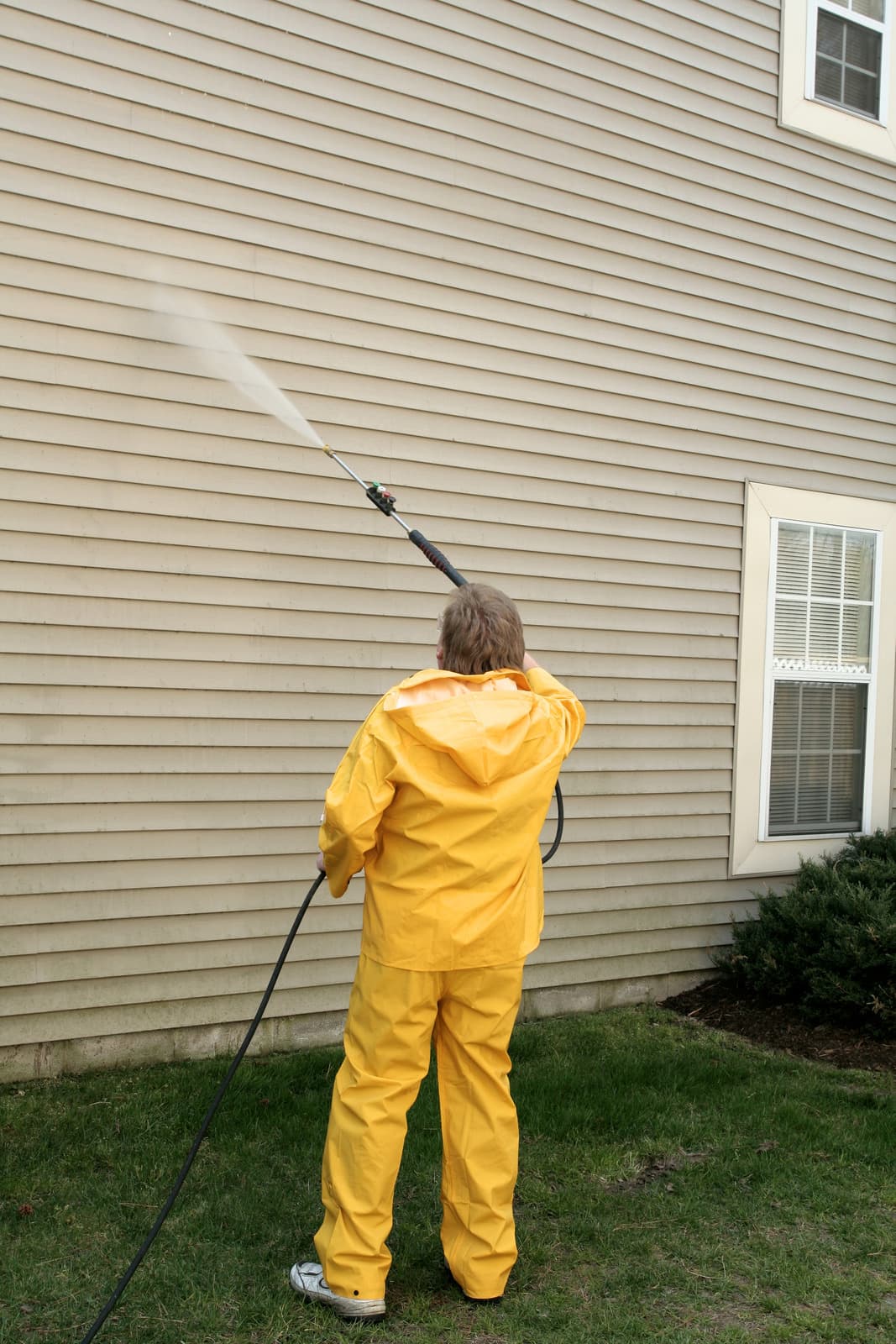 Technician in yellow waterproof gear soft washing beige vinyl house siding